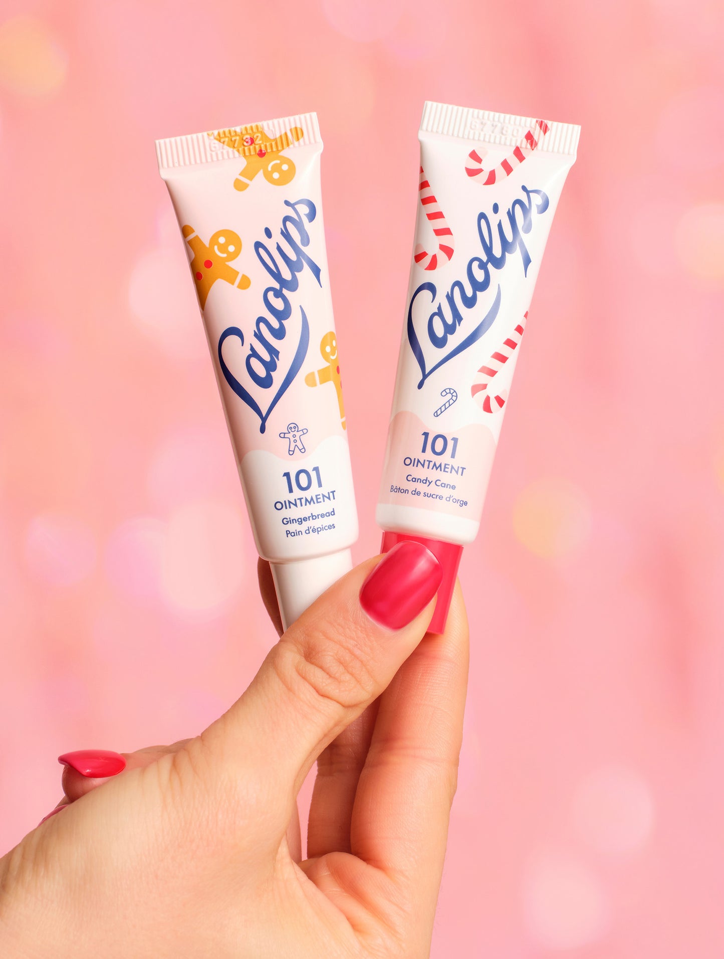 The limited edition Lanolips Holiday Gingerbread House contains the 101 Ointment Multi-Balms in Gingerbread and Candy Cane lip balm tubes held by a hand against a pink background. 